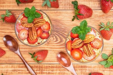 Birkaç bardak yulaf ezmesi, bal ve üzerinde nane yaprağı olan fındıklı çilekli parfe. Bir tarafta bütün çilekler ve ahşap bir masada tahta kaşıklar. en üstteki görünüm
