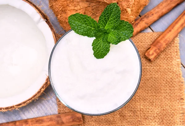 coconut smoothie with a mint leaf on top, on the side whole coconuts and cinnamon sticks on a table of gray wooden planks. rustic concept. in top view