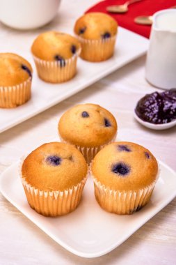 Beyaz tabakta küçük yaban mersinli kekler, muffinlerin bulanık arka planı, böğürtlen reçeli ve beyaz mermer yüzeyde kahve kreması. Süslü. portre görünümü