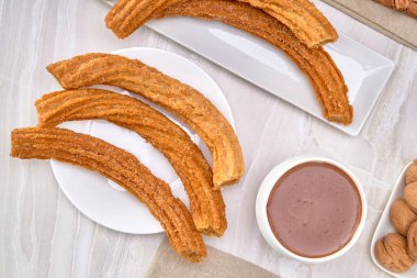 Beyaz bir tabakta şeker ve tarçınla kaplı İspanyol churro 'larına mermer bir zemin ve beyaz bir zemin üzerinde beyaz bir bardakta İspanyol çikolatası eşlik ediyor. en üstteki görünüm