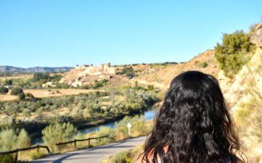 Uzun saçlı, esmer turist Zorita de los Canes kasabasını ve kalesini uzaktan izliyor. İkisi arasında Tajo nehri.