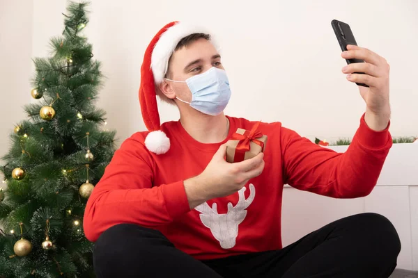 Christmas online greetings in a mask. Man in santa claus hat and medical mask talking on the phone near christmas tree.