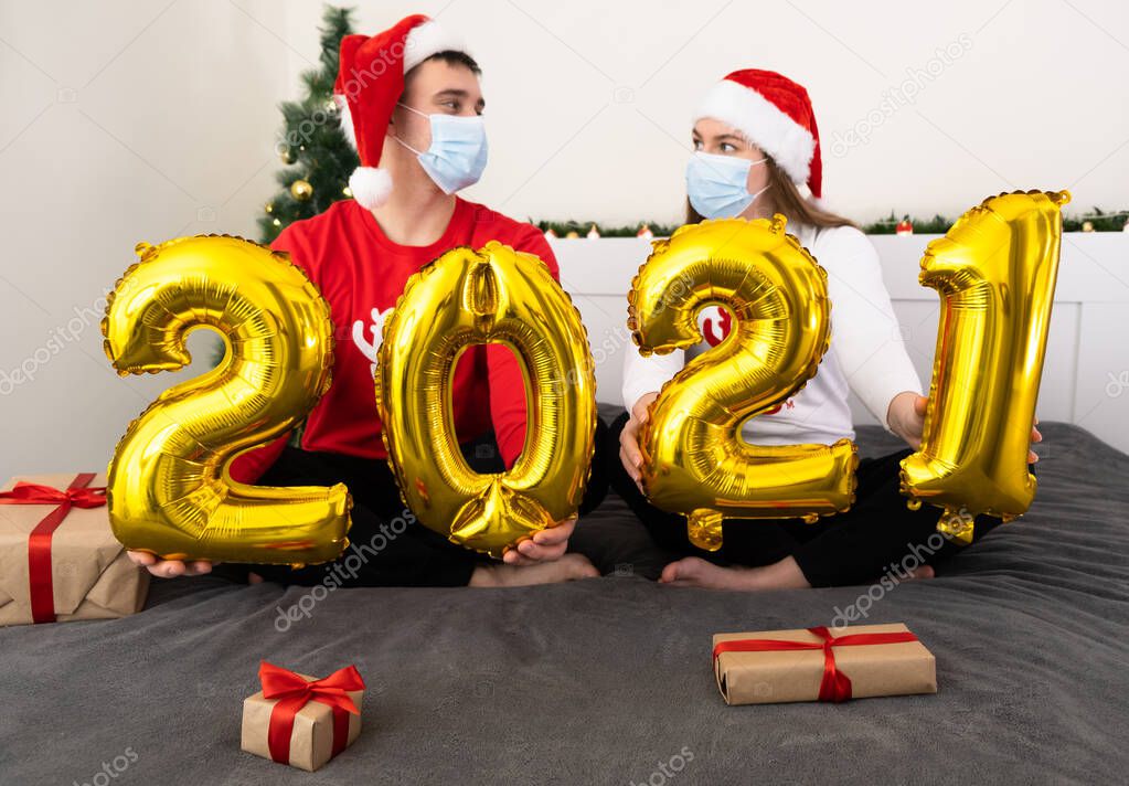 A young couple in masks and santa claus hats in a homely atmosphere holds golden balloons number 2021 in their hands. Christmas and coronavirus.