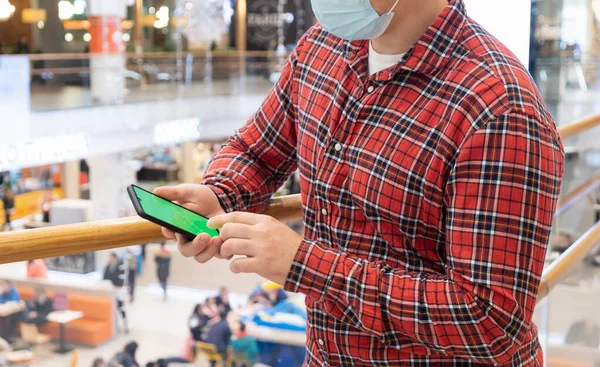 A young man in a medical mask in a shopping center looks into his smartphone. Covid-19.