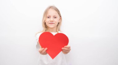 Beyaz tişörtlü küçük bir kız beyaz arka planda kırmızı bir kalp tutuyor. Sevgililer Günü için tebrik kartı, Anneler Günü, Babalar Günü.