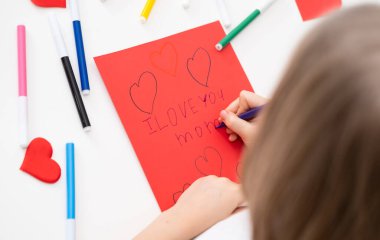 Çocuk Anneler Günü bayramı için bir kartpostal çizer. Kız kırmızı kağıda yazıyor. Seni seviyorum anne..