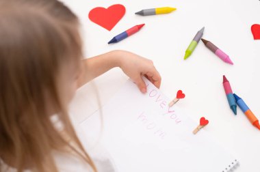 Çocuk Anneler Günü bayramı için bir kartpostal çizer. Kız beyaz kağıda yazıyor, seni seviyorum anne..