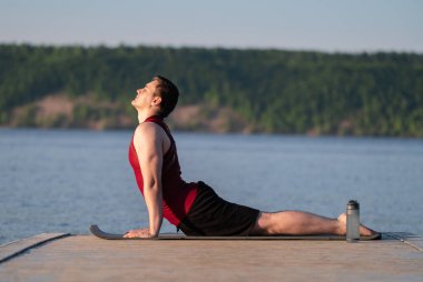 Genç bir adam nehir kıyısında yoga yapıyor. Doğada kilo verme ve spor yapma kavramı.