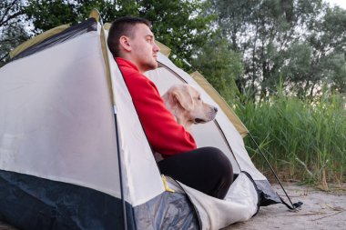 Gülümseyen genç adam köpeğiyle çadırda oturuyor. Kamp alanındaki Golden Retriever 'lı adam. Hayvanlarla seyahat kavramı.