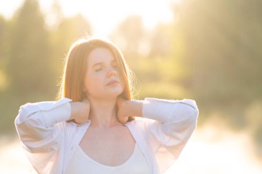 A beautiful red-haired girl in a white shirt meets the summer dawn in nature. The concept of happiness and spiritual freedom.