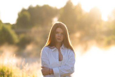 A beautiful red-haired girl in a white shirt meets the summer dawn in nature. The concept of happiness and spiritual freedom.