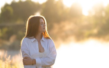 A beautiful red-haired girl in a white shirt meets the summer dawn in nature. The concept of happiness and spiritual freedom.