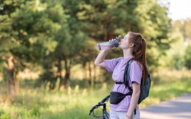 Redhead girl rides a bicycle in a beautiful summer forest. Woman cyclist drinks water from a reusable bottle