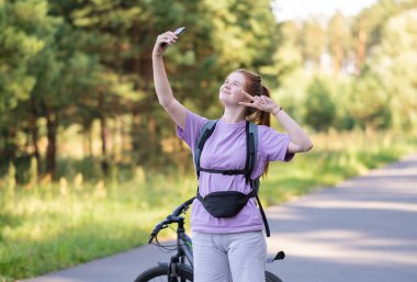 Redhead girl rides a bicycle in a beautiful summer forest. Woman cyclist looks at the phone in the park.