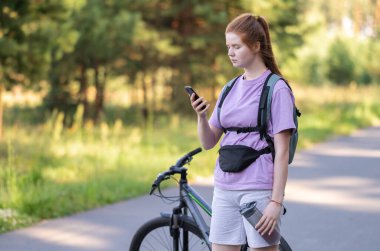 Redhead girl rides a bicycle in a beautiful summer forest. Woman cyclist looks at the phone in the park.