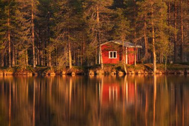 A small and cozy wilderness cabin with reflection during sunset by the summery lakeside in Finnish countryside in the middle of taiga forest, Northern Europe. clipart