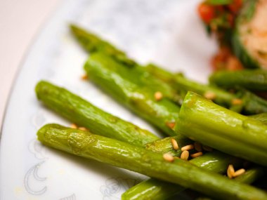 Beyaz çiçekli bir tabakta sunulan susam tohumuyla pişirilmiş yeşil kuşkonmaz, dengeli bir diyet için vitamin ve lifle dolu sağlıklı bir vegan mezesi sunuyor..