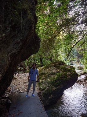 Erkek bir yürüyüşçünün, doğa yürüyüşü sırasında kayalık bir kanyonda, büyük bir kayanın ve akan bir nehrin yanında beton bir yolda durduğu tüm vücut görüntüsü.