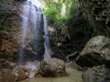 Şelalenin geniş açılı görüntüsü açık bir havuza akıyor. Büyük kayalar ve güneşli bir orman kanyonundaki yeşil ağaçlarla çevrili.