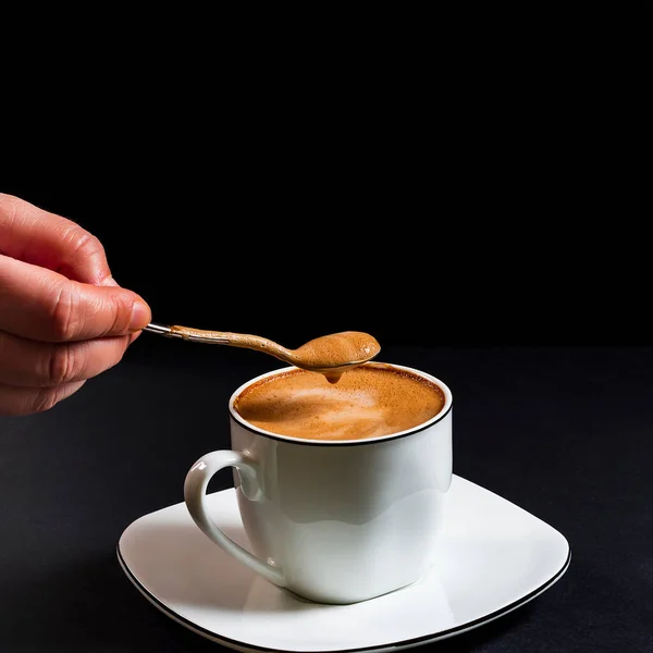 Person picking up a latte coffee spoon.This is a square-format photo taken on a black background under artificial light in a studio.