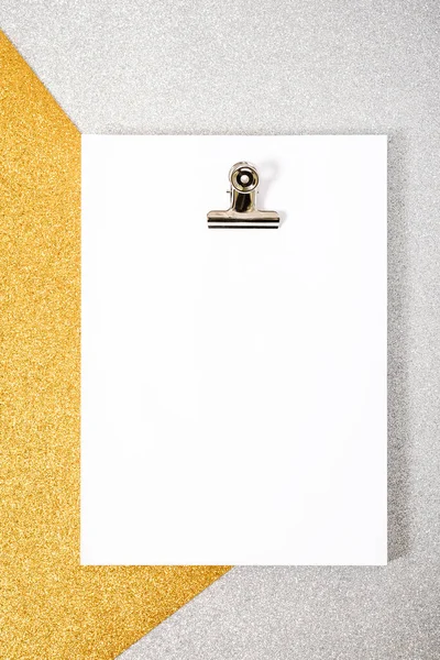 White foil packet and paper clip on a silver and gold textured background. The photo has a copy space, it is taken from an overhead point of view and is in vertical format.