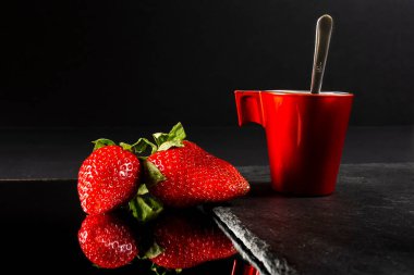 Two fresh red strawberries reflected on a mirror and a red cup on a slate base.The photograph is a still life taken with artificial light and in horizontal format.