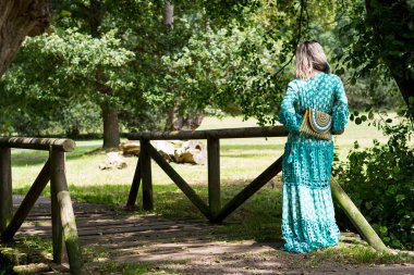 İspanyol sarışın kadın, Asturias 'ta bir parktaki ahşap bir köprüde sırtüstü poz vererek çantasını ve uzun yeşil elbisesini gösteriyor. Fotoğrafın güzel bir manzarası var ve yatay olarak çekiliyor..