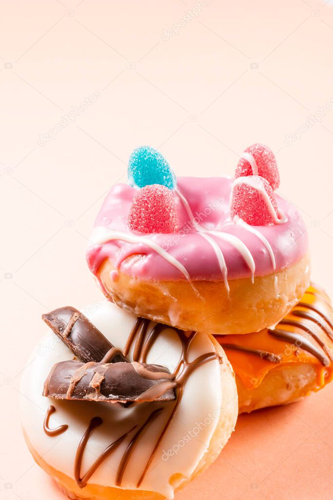 Photograph of 3 donuts decorated with jelly beans and drawn with chocolate.The photo is taken in vertical format on a cream colored background.The photo has copy space.