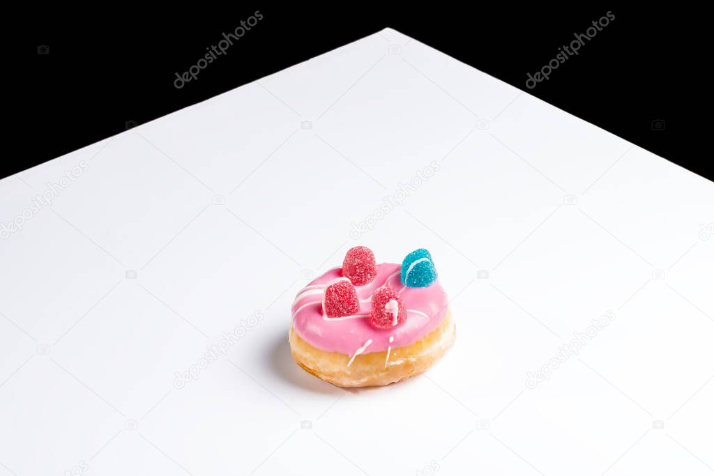 Photo of a pink donuts painted with white chocolate and decorated with jelly beans.The photograph is taken in horizontal format on a white table and has a copy space.