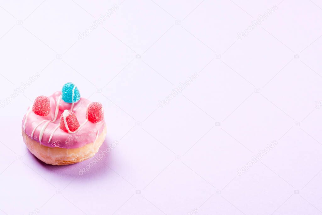 Photograph of 1 pink donuts decorated with jelly beans and drawn with white chocolate.The photo is taken in horizontal format on a lilac background and has copy space.