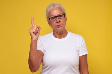 Gözlüklü orta yaşlı bir kadın parmağıyla işaret ederek ciddi bir ifade takınıyor.