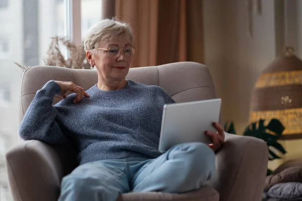 Senior kvinna med surfplatta vilar på soffa nära fönstret läser något på internet. — Stockfoto