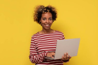 Genç Afrikalı, kıvırcık saçlı, dizüstü bilgisayar kullanan ve sarı üzerine izole edilmiş gülümseyen havalı bir bayan.