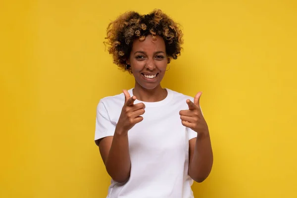 あなたを指差しながらカメラを見ている幸せな若いアフリカの女性 黄色の壁にスタジオショット — ストック写真
