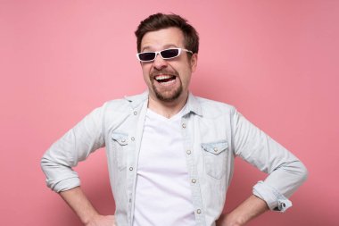 Güneş gözlüklü genç yakışıklı adam pembe duvarda gülümsüyor..