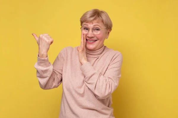 Зрелая белая женщина, указывающая указательным пальцем в сторону, шепчет тайну за рукой — стоковое фото