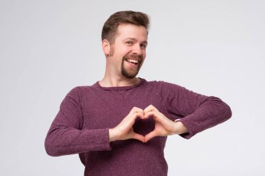 Sakallı genç adam, aşkla gülümsüyor, elleriyle kalp sembolü yapıyor..