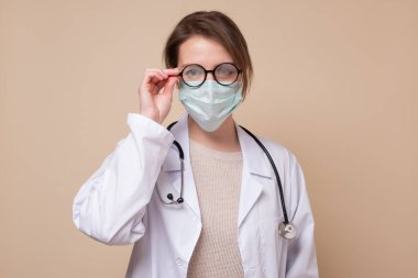 Tıbbi maskeli ve gözlüklü genç bayan doktor kameraya bakıyor..