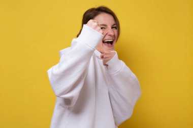 Sarı arka planda izole bir şekilde duran enerjik genç kadın yumruklarını sıkıyor. Stüdyo çekimi