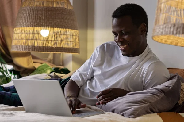 Молодий позитивний усміхнений африканський чоловік друкує на ноутбуці вдома — стокове фото