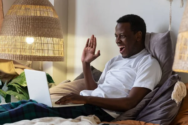 Black man with laptop resting in bed greeting waving hand. — Stock Photo, Image