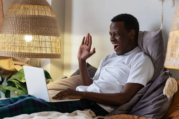 Black man with laptop resting in bed greeting waving hand. Royalty Free Stock Photos
