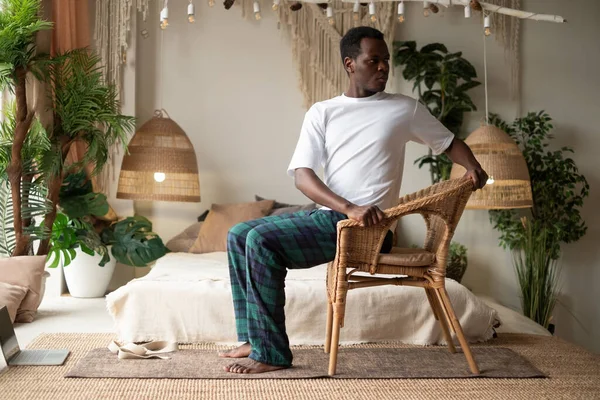 Sporty young african man practicing yoga using chair, doing Revolved Chair Pose, Parivrtta Utkatasana Stock Image