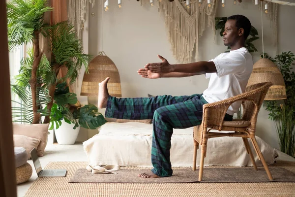 African man using chair at his living room at home doing yoga asana for beginners. Stock Picture