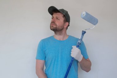Mavi tişörtlü, kepli ve eldivenli genç ressam boya silindirli bir duvarı boyuyor..