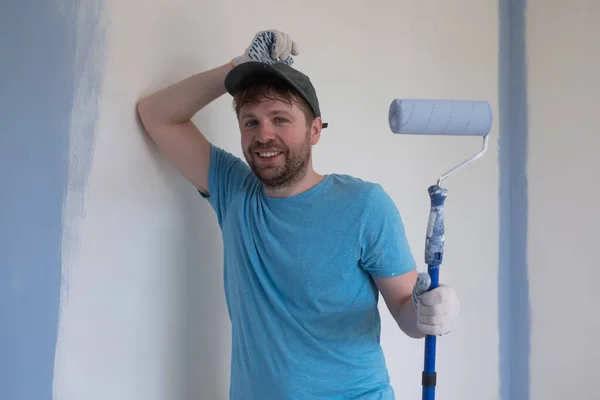 Joven pintor en camiseta azul, gorra y guantes pintando una pared con rodillo de pintura. — Foto de Stock