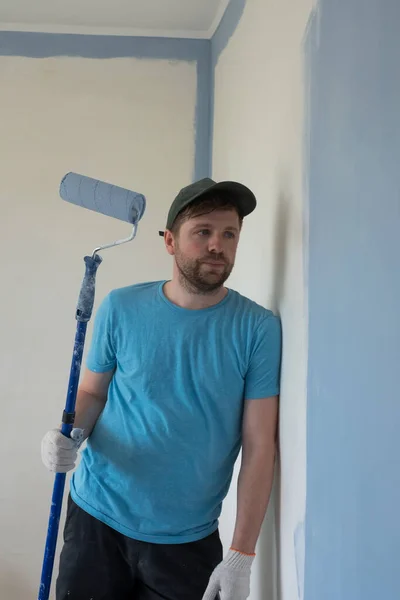 Joven pintor en camiseta azul, gorra y guantes pintando una pared con rodillo de pintura. — Foto de Stock