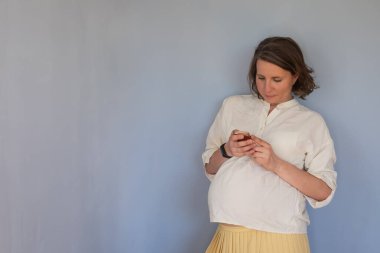 Mavi duvarda cep telefonu gülümsemesi kullanan hamile bir kadın..