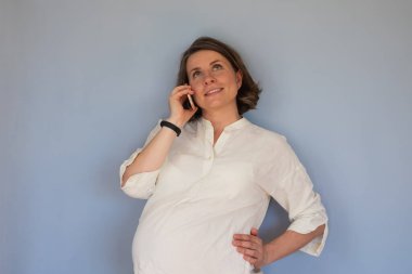 Mavi duvarda cep telefonu gülümsemesi kullanan hamile bir kadın..