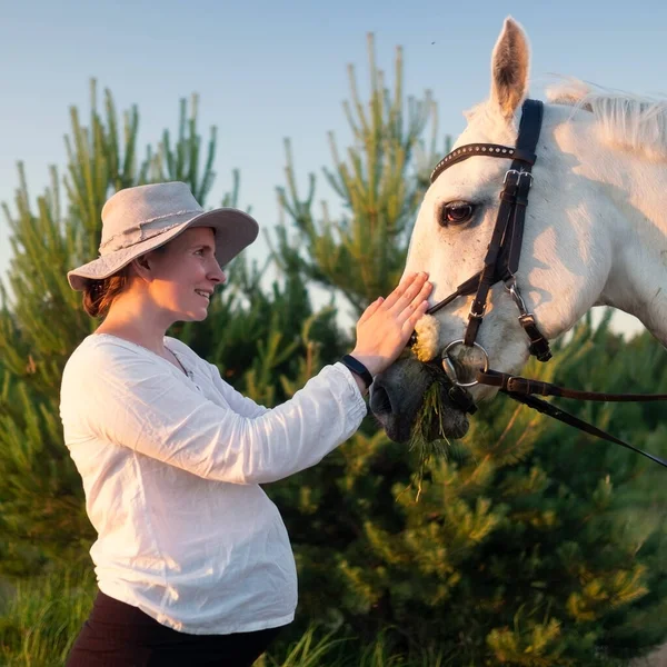 Jovem grávida alimentando um cavalo branco no campo — Fotografia de Stock